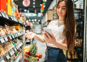 Une femme dans un supermarché, utilisant son téléphone et portant un panier rempli de fruits, symbolisant l'identification des aliments déclencheurs pour la rectocolite hémorragique (RCH), la maladie de Crohn et autres MICI.